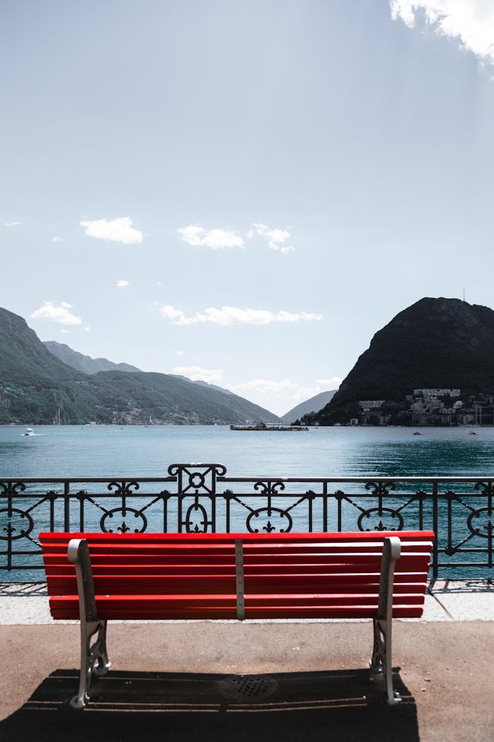 Vibrant red bench by the tranquil Lake Lugano offering picturesque mountain views.